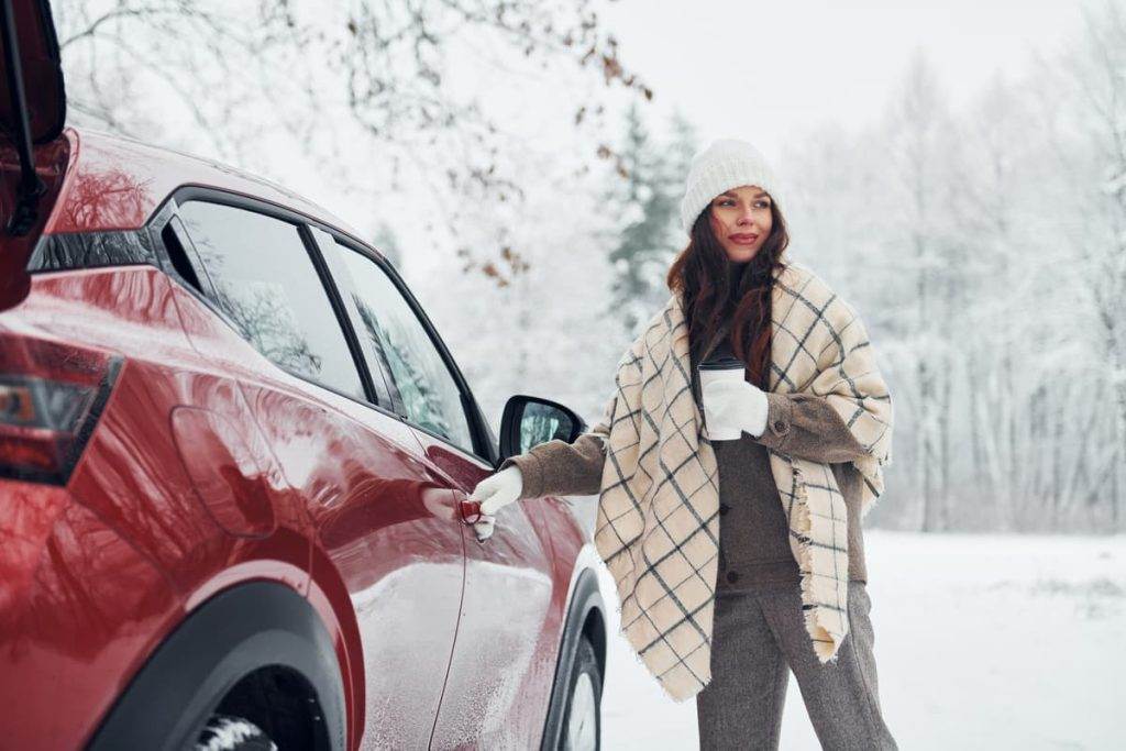 Femme en hiver ouvrant la porte de sa voiture rouge enneigée, tenant un café chaud